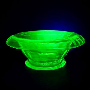 Puede incluir: Un cuenco de cristal de color verde neón brillante y vibrante, con un borde ancho y plano con detalles lineales grabados y una base circular. El cuenco emite una luz brillante e intensa sobre un fondo oscuro, resaltando su estructura translúcida.