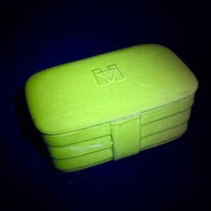 May include: A lime green rectangular box with a textured surface and a small, embossed logo on the lid. The box has a hinged design with horizontal lines and a clasp. The background is a dark blue.