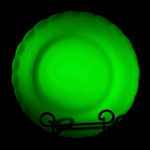 May include: A glowing green glass plate with a ruffled edge. The plate is round and sits on a black metal stand, with a dark background. The plate's color is a vibrant, bright green.