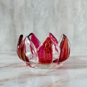 May include: A clear glass votive candle holder with a flower petal design. The petals are a vibrant red and pink color, with clear glass accents. The candle holder is sitting on a marble surface.