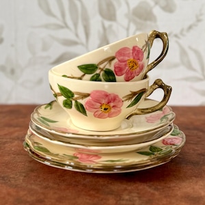 May include: Cream-colored teacups and saucers with pink rose floral designs. The teacups are stacked, and the saucers are arranged below. The handles of the teacups have a branch-like design. The set is a vintage style.