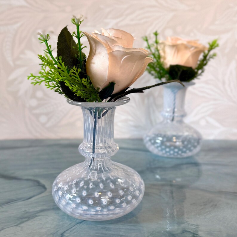 Op de afbeelding: Twee heldere glazen vaasjes met een textuur van belletjes. Elke vaas bevat een perzikroos met groene bladeren. De vaasjes staan op een lichtblauwe ondergrond, wat zorgt voor een delicate en elegante bloemstuk.