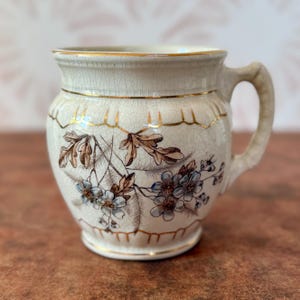 May include: Cream-colored ceramic mug with a floral design featuring blue and brown flowers and leaves. The mug has a gold rim and handle, and a decorative gold band. The mug has a crackled finish.