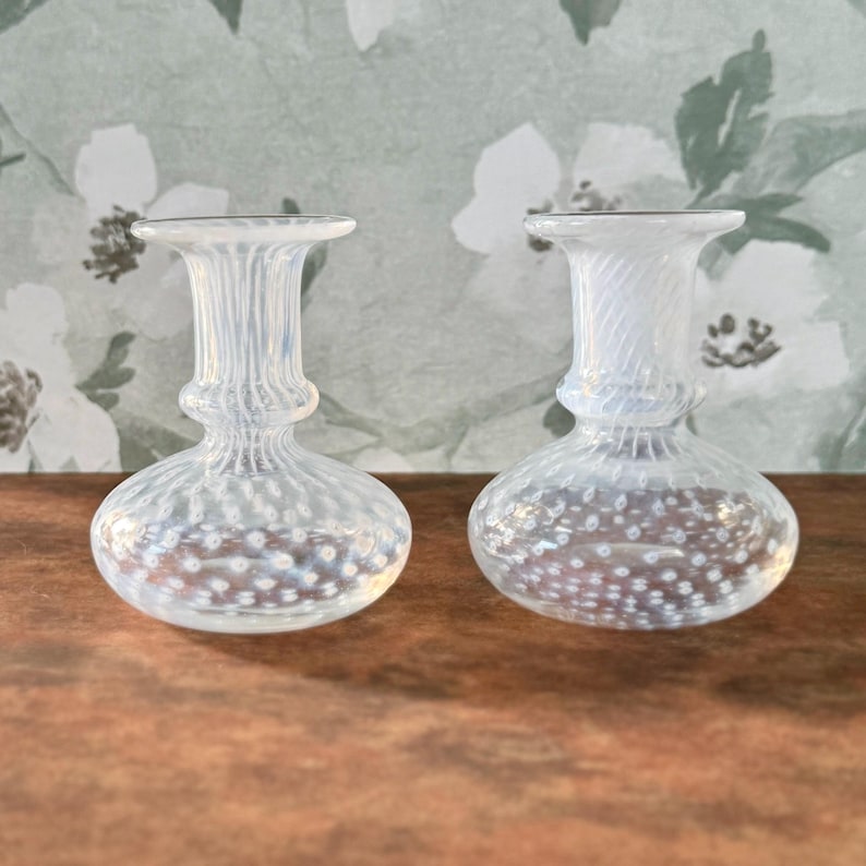 Op de afbeelding: Twee kleine, heldere glazen vazen met een bolvormige basis en een uitlopende bovenkant. De vazen hebben een textuur met stippen en worden gepresenteerd op een houten oppervlak. De achtergrond is een bloemenbehang.