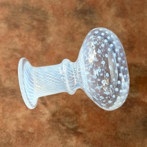 Op de afbeelding: Een heldere glazen flesstopper met een bolvormige bovenkant en een cilindrische basis. De bovenkant is bedekt met kleine, witte, gelijkmatig verdeelde stippen. De basis heeft een textuur, geribbeld ontwerp. De stopper is ontworpen om in een fles te passen.