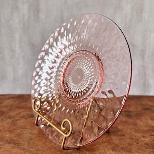 May include: A pink glass plate with a diamond pattern and a decorative center design. The plate is displayed on a gold-colored metal stand, showcasing its intricate details and vintage aesthetic. The plate's rim has a slightly darker pink hue.