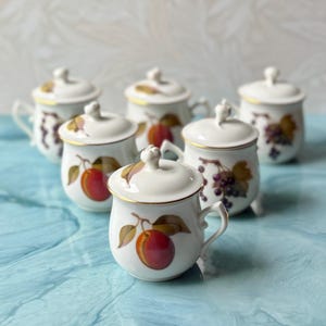 May include: A set of five white porcelain lidded pots, each featuring a gold-trimmed rim and handle. The pots are decorated with colorful botanical illustrations of peaches and grapes, with matching lids topped by small, rounded finial handles.