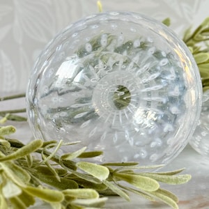 Op de afbeelding: Een heldere glazen decoratieve bol met een textuuroppervlak en kleine witte bellen. De bol heeft een gedetailleerde, spiraalvormige basis. Groene bladsprietjes staan op de voorgrond.