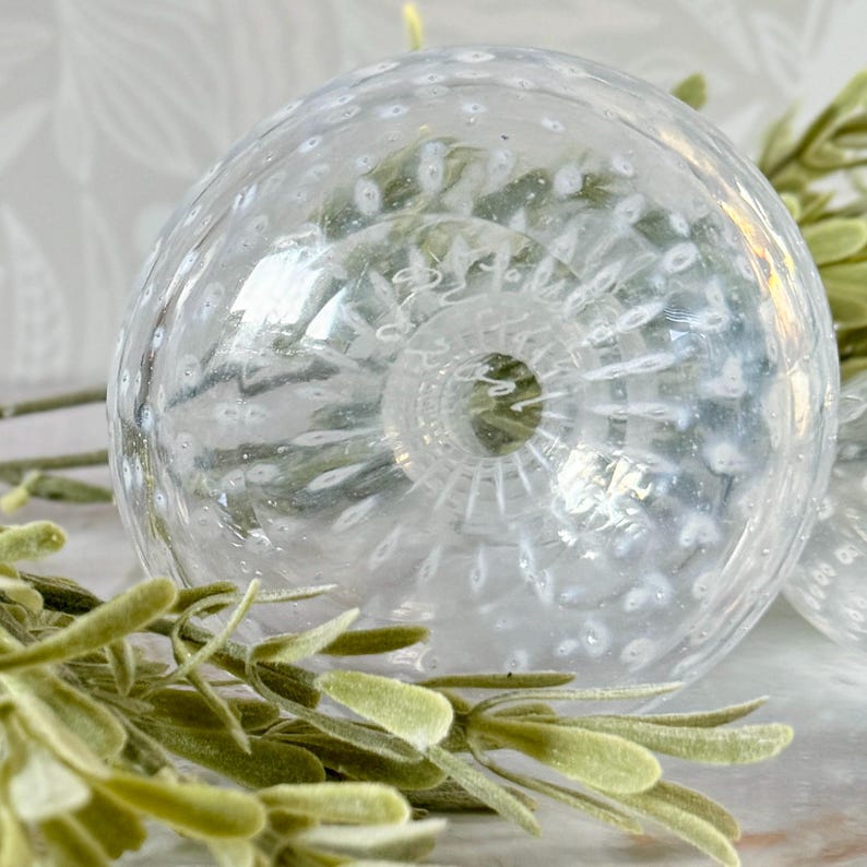 Op de afbeelding: Een heldere glazen decoratieve bol met een textuuroppervlak en kleine witte bellen. De bol heeft een gedetailleerde, spiraalvormige basis. Groene bladsprietjes staan op de voorgrond.