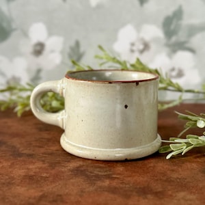 May include: A small, speckled beige ceramic mug with a rounded handle and a slightly flared base. The rim is a darker brown color. The mug is approximately 3 inches tall and is suitable for coffee or tea.