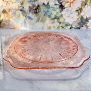 May include: A pink, square glass serving plate with a raised floral design in the center and along the edges. The plate has a scalloped edge and is made of clear glass. The plate is likely vintage and suitable for serving appetizers or desserts.