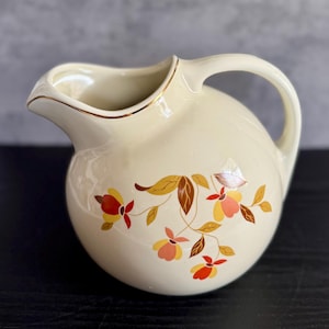 May include: Cream-colored ceramic pitcher with a rounded body and a curved handle. The pitcher features a floral design in shades of yellow, orange, and brown. The rim is trimmed with a gold accent. A decorative piece for serving beverages.