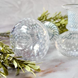 Op de afbeelding: Decoratieve objecten van helder glas, waaronder een bol en een kleine vaas. De bol heeft een gespikkeld patroon en de vaas heeft een gedraaid ontwerp. Groen voegt een natuurlijk accent toe. De objecten staan op een marmeren oppervlak.