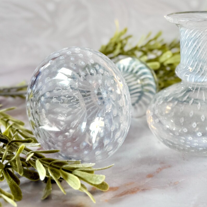 Op de afbeelding: Decoratieve objecten van helder glas, waaronder een bol en een kleine vaas. De bol heeft een gespikkeld patroon en de vaas heeft een gedraaid ontwerp. Groen voegt een natuurlijk accent toe. De objecten staan op een marmeren oppervlak.