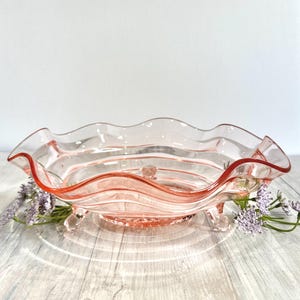 May include: A pink glass footed bowl with a scalloped edge. The bowl has a wavy design and is perfect for serving fruit or snacks.