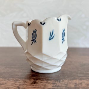 以下が含まれることがあります： 取っ手とスカラップリムが付いた白いクリーマー。クリーマーは、下半分に幾何学的なダイヤモンド模様があり、青い花の模様で装飾されています。クリームやミルクを提供するのに最適なクラシックな作品です。