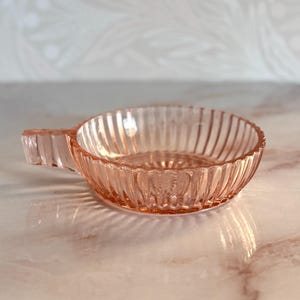 May include: A small, round, pink glass dish featuring a ribbed exterior pattern and a single, short, rectangular handle extending from one side. The glass is translucent with a smooth, polished finish, showcasing vertical ridges along the bowl.