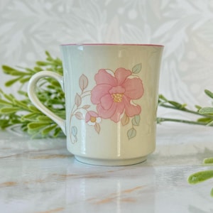 May include: A ceramic mug with a floral design. The mug is off-white with a pink rim and handle. A large pink flower with green leaves is the central design element. The mug is approximately 4 inches tall.