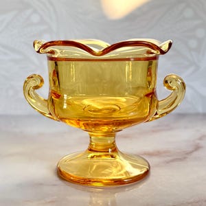 May include: A vintage amber-colored glass bowl with a scalloped rim and two curved handles. The bowl sits on a pedestal base. The glass has a glossy finish, reflecting light. This decorative piece is likely from the mid-century era.