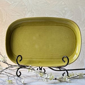 Könnte beinhalten: Eine ovale Servierplatte aus Keramik mit einer glänzenden, chartreuse-grünen Glasur und einem glatten, leicht erhöhten Rand. Die Platte wird auf einem schwarzen Metallständer mit Schnörkeln präsentiert und zeigt ihre gleichmäßige Farbe.