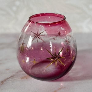May include: A clear, round glass vase with a deep pink colour and gold starburst designs. The vase has a wide opening and a rounded body, ideal for displaying small flowers or decorative items.