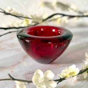May include: A small, ruby-red glass bowl with a rounded shape. The bowl has a dark rim and a smooth, glossy surface. White flowers and branches are in the background, adding a touch of elegance. This decorative bowl is suitable for holding small items.