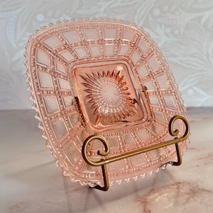 May include: A square pink depression glass plate featuring a central starburst pattern surrounded by a grid of textured squares and a scalloped edge. The plate is displayed upright on a gold-toned metal wire stand with two curved supports.