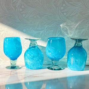 May include: Four hand-blown, turquoise-colored glass goblets. Each goblet has a unique shape and design, with a textured, swirled appearance. The stems and bases are clear, and the bowls are a vibrant blue.