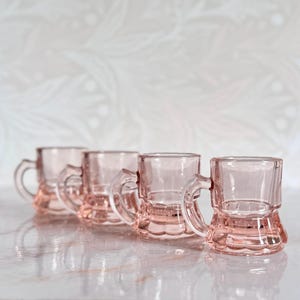 May include: A set of four small, translucent pink glass mugs arranged in a row. Each mug features a sturdy handle, a smooth cylindrical upper body, and a decorative scalloped base design. The glass has a light, rosy tint and a polished finish.