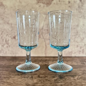 May include: Two clear, stemmed glasses with a light blue base and rim. The glasses have a tall, cylindrical shape and are set on a wooden surface. The base of each glass has a fan-like design.