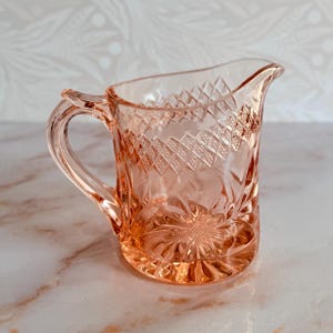 May include: A translucent pink glass creamer pitcher featuring a textured diamond pattern around the upper rim and a pressed floral starburst design on the lower body. The vessel includes a curved handle and a small pouring spout.