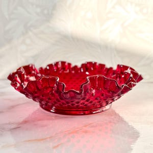 Puede incluir: Un cuenco de vidrio rojo rubí con borde ondulado. El cuenco tiene un patrón de clavos y se asienta sobre una pequeña base. El borde ondulado tiene un ligero ribete dorado. Este cuenco vintage es ideal para servir o exhibir.