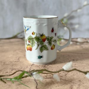May include: White ceramic mug with a gold rim and handle. The mug is decorated with a colorful design of strawberries, green leaves, butterflies, and small insects. The mug has a slightly textured surface.