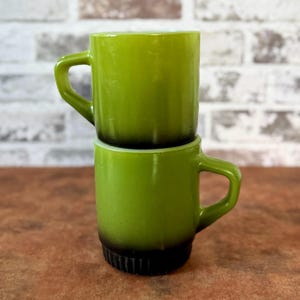 May include: Two vintage green mugs stacked on top of each other. The mugs have a rounded shape with a handle and a dark brown base. The mugs are made of a glossy material and are set against a brick wall.