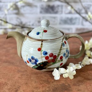 May include: A ceramic teapot with a lid, spout, and handle. The teapot is white with a textured surface and is decorated with blue and red floral designs. The spout and handle are a light brown colour.