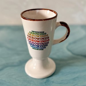 May include: A white ceramic mug with a brown rim and handle. The mug features the word "CAROWINDS" in a rainbow-colored, stacked design. The mug has a pedestal base.