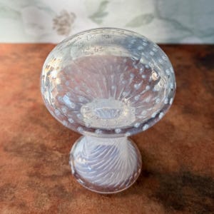 Op de afbeelding: Een decoratief, paddenstoelvormig object van helder glas. De bovenkant is een afgeronde koepel met kleine luchtbellen. De steel heeft een wervelontwerp. Het object staat op een bruin oppervlak.