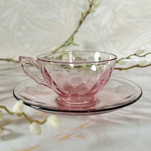 Könnte beinhalten: Ein rosa Teetassen- und Untertassen-Set aus Glas. Die Teetasse hat ein Diamantmuster und einen Henkel. Die Untertasse ist rund mit leicht erhöhtem Rand. Das Set besteht aus transparentem rosa Glas.