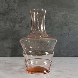 May include: A clear glass decanter with a pink hue and a textured, rounded body. The decanter has a long, narrow neck.