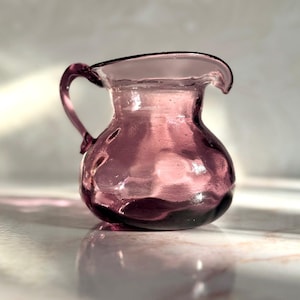 May include: A small, antique-style pitcher made of translucent purple glass. The pitcher has a rounded body, a curved handle, and a spout. The glass has a slightly textured surface, and the pitcher is sitting on a marble surface.