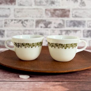 May include: Two white ceramic teacups with a green floral pattern around the rim. The cups have a rounded shape and small handles. They are placed on a round, wooden tray against a brick wall background.