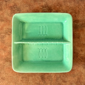 Puede incluir: Un plato cuadrado de cerámica verde menta con un divisor central de borde dentado. El plato tiene dos compartimentos simétricos, cada uno con un diseño en relieve de tres bucles cursivos centrado en la base de la superficie interior.