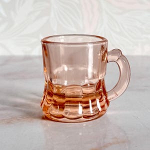 May include: A small, translucent pink glass mug featuring a smooth upper rim and a base with vertical fluted panels. The mug has a curved handle on the right side and a clear, polished finish that reflects light across its rounded, cylindrical body.