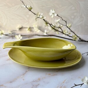 May include: A chartreuse ceramic gravy boat with a teardrop-shaped bowl and a matching oval underplate. The glaze features a subtle crackle texture throughout the surface, and the vessel includes a pointed pouring spout and a handle.