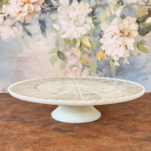 May include: A white, vintage-style cake stand with a scalloped edge and a detailed, starburst-like pattern on the top. The stand has a short, rounded base and is made of opaque glass. The background features a floral painting.