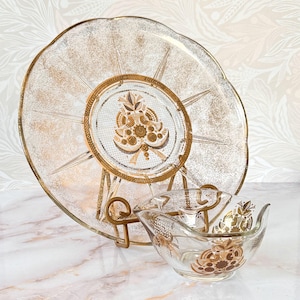May include: A vintage glass serving set with a large, round plate and a small bowl. The plate and bowl have gold accents and a decorative design. The set is displayed on a gold metal stand, perfect for serving snacks or appetizers.
