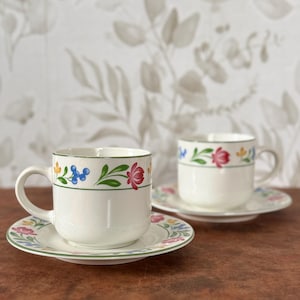May include: Two white ceramic teacups and saucers with floral designs. The cups and saucers have a green border and are decorated with colorful flowers and leaves. The teacups are on a brown surface.