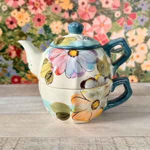 May include: A ceramic teapot and teacup set. The set is painted with large, colorful flowers in shades of pink, orange, and blue. The teapot has a blue lid and handle, and the teacup is stacked underneath. The background is a floral pattern.