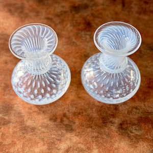 Op de afbeelding: Twee heldere glazen parfumflesjes. De flesjes hebben een ronde basis met een textuurpatroon en een uitlopende bovenkant met een wervelontwerp. De flesjes staan op een bruin oppervlak.