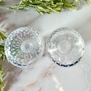 Op de afbeelding: Twee heldere glazen kandelaars met een bubbelontwerp. De kandelaars hebben een ronde vorm met een gedetailleerde basis en zijn omgeven door witte stippen. De kandelaars staan op een marmeren oppervlak.
